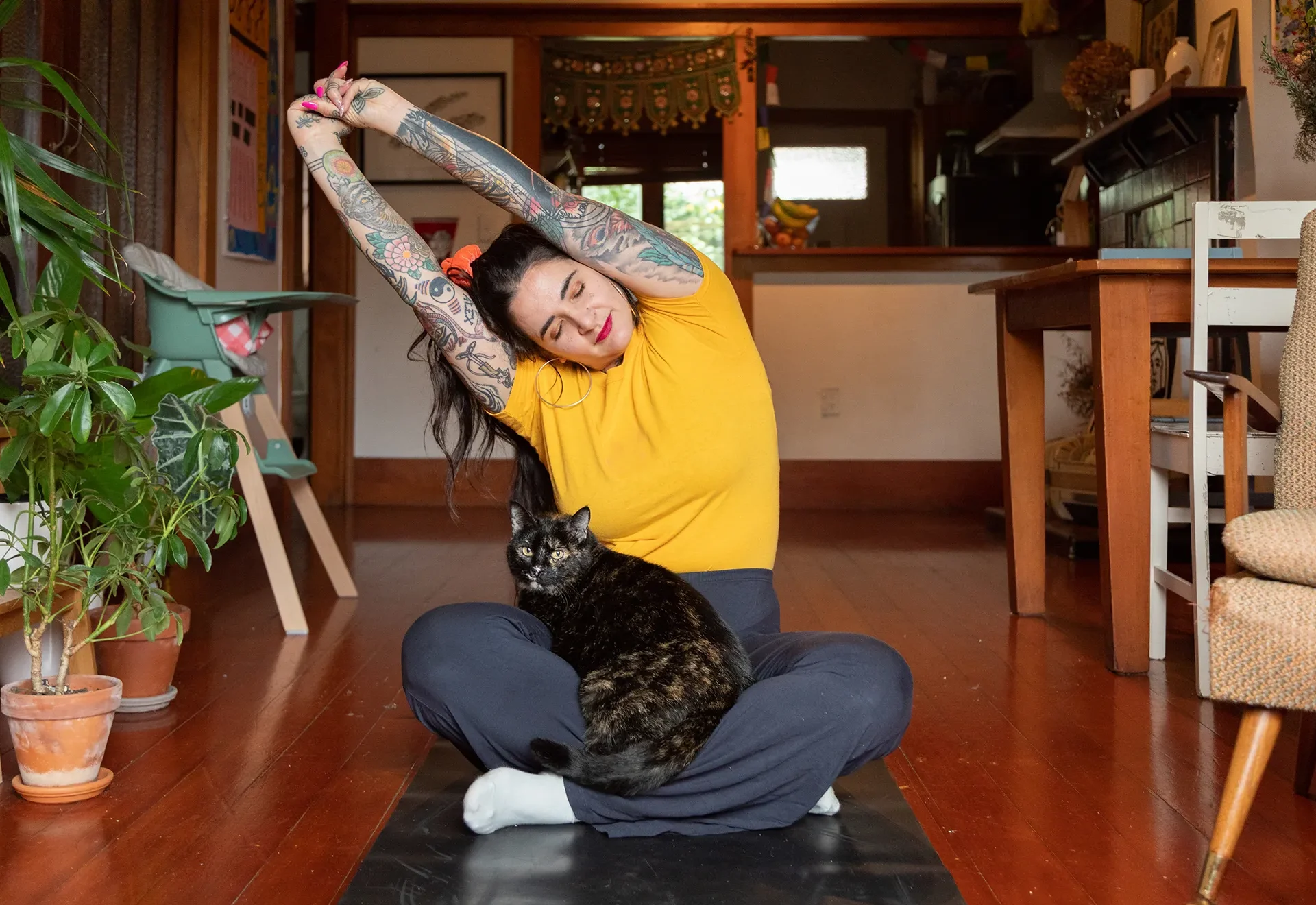 Woman sitting cross legged on a yoga mat indoors stretching her arms up in the air while a cat sits on her lap